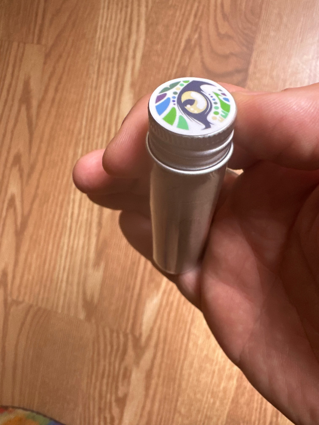 Hand holding a small cylindrical container with ashes from Tsunu bark and a colorful cap on a wooden surface